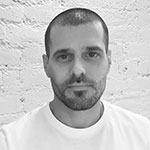 A black and white portrait of a man with a shaved head and beard, wearing a white t-shirt against a white brick wall background.